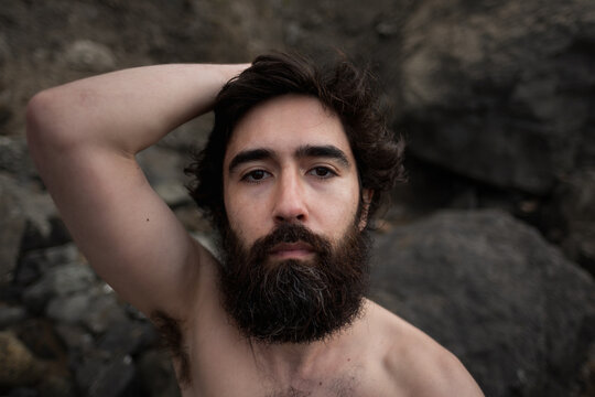 A scruffy, bearded man with messy hair sits on a rocky beach and gazes at the camera. He lifts up his arm, showing his skinny physique.