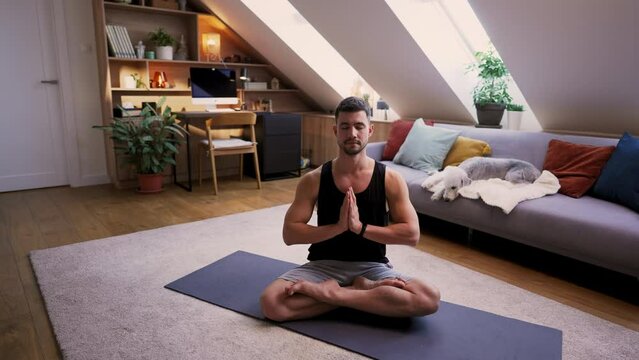 Calm man sitting at lotus position with closed eyes and having practice yoga at home while his dog laying at the sofa