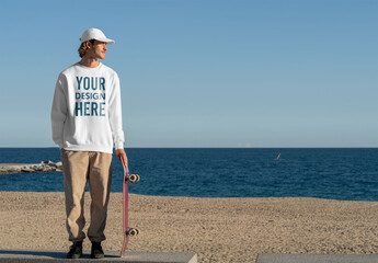 Mockup of man wearing customizable sweatshirt with skateboard by sea