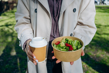 A woman holds a takeaway coffee and a bowl of vibrant, mixed green salad with fresh strawberries, ready for a delightful meal in a sunlit park