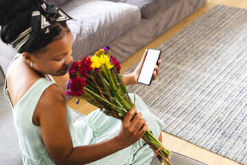 African American woman holding flowers, looking at smartphone on a video call date with copy space