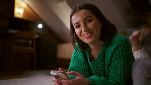 Young woman laying down on the living room floor and using a smartphone with happy face