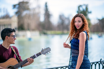 Two young adults enjoying a music session by a lake in a sunny park, one playing the guitar...