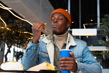 African American man in a denim jacket and orange beanie savors his meal, with a soda cup in hand, in the vibrant ambiance of a contemporary cafe. 