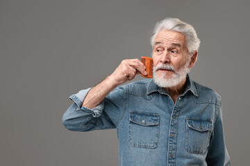 Senior man combing beard on grey background, space for text