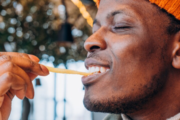 Close-Up of a Smiling African American Man Enjoying French Fries in a Cozy Setting. Smiling man enjoying a French fry, the joy in the simple pleasure of eating captured against a bokeh light