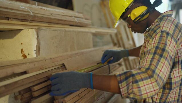 Side view of African American carpenter or woodworker or charpentier man use measuring tape to measure wood stick to prepare wood work in workplace.