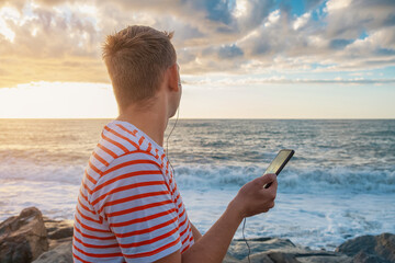 Back view ofCaucasian young man listening to musc with headphones using mobile phone on the sea...