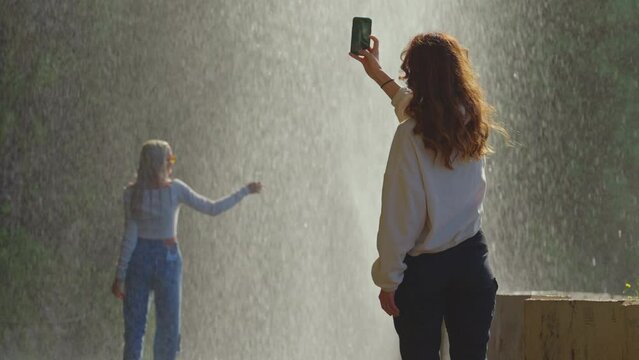 Girl taking pictures on smartphone of another happy woman in waterfall drops having fun enjoying freedom with arms raised feeling carefree tropical vacation