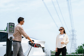 Young couple recharge EV car battery at charging station connected to power grid tower electrical...