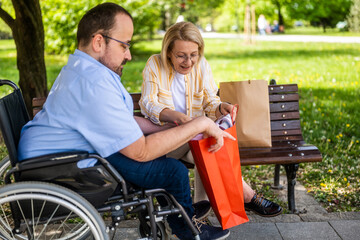Man in wheelchair is spending time with his mother in park. They are talking after shopping. - 785111407