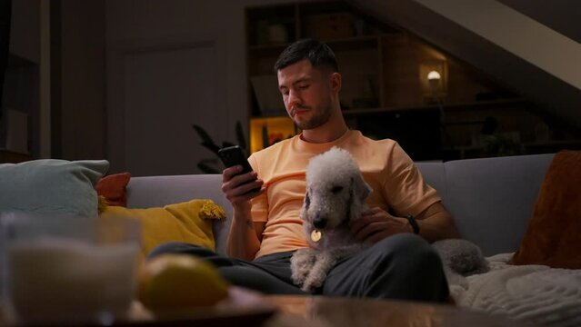 Young man using mobile phone in his room, cuddling with his dog