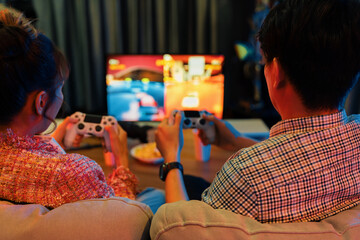 Couple gamer with joysticks playing fighting video game together on tv screen, getting challenge level at back side view, sitting sofa at neon light color living room at modern comfy home. Infobahn.