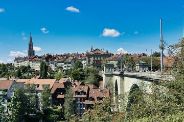 Cityscape of Bern with red-tiled buildings and lush trees against a blue sky in the background.