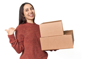 Caucasian young woman with moving boxes on studio background points with thumb finger away, laughing and carefree.