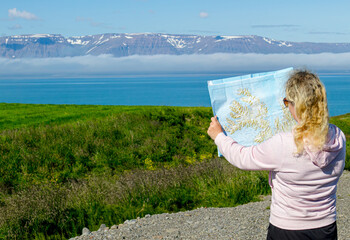 Young woman tourist is looking at the Iceland Westfjords map in idyllic nature with Westfjord snow capped mountains on background and fjord.