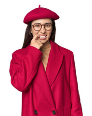 Stylish young Caucasian woman in red trench coat and beret on studio background biting fingernails,...