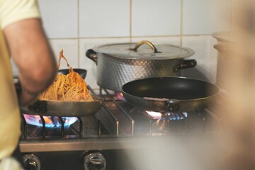 Nice shot of a male preparing spaghetti in a frypan, with other pots on the cooker with flames on
