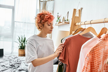attractive red haired extravagant person picking up stylish clothes near rack at home, leisure time