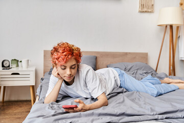 beautiful relaxing queer person in homewear with red hair lying on bed and using her smartphone