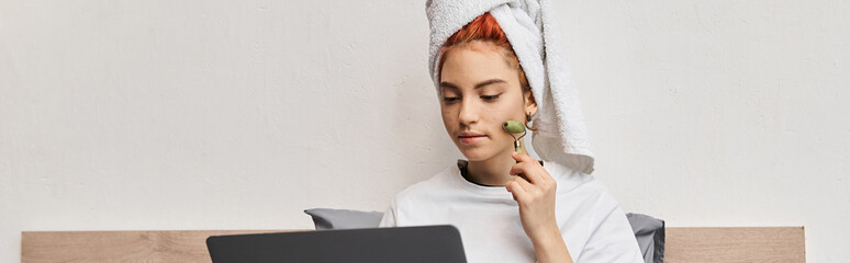 appealing queer person in homewear using face roller while relaxing on her bed with laptop, banner