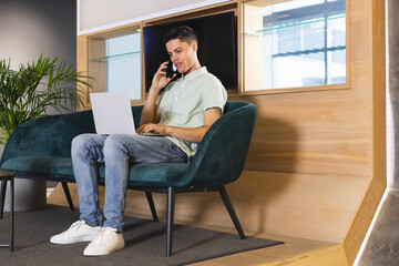 A biracial man talking on phone, holding laptop on his lap in a modern business office