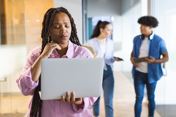 Biracial woman holding laptop, thinking, colleagues walking behind in a modern business office