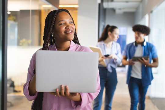 A young biracial woman holding a laptop, colleagues chatting behind in a modern business office