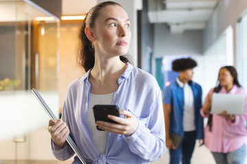 Caucasian female holding smartphone, walking in hallway in a modern business office