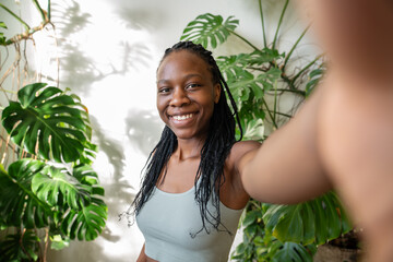 Joyful African American blogger girl make selfie on smartphone while standing in sunny room with...