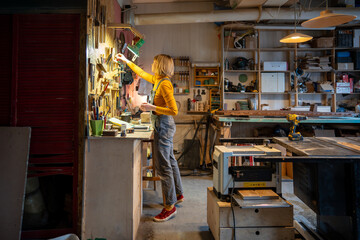 Focused on workflow in carpentry shop craftswoman carpenter in denim picks up tools to produce handmade wooden furniture. Small artisan business. Concentrated female cabinetmaker busy joinery workshop