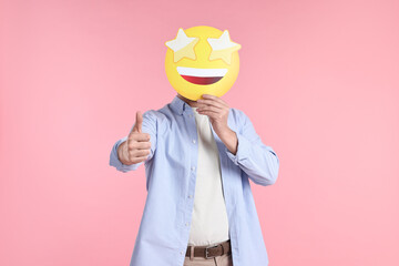 Man holding emoticon with stars instead of eyes and showing thumb up on pink background