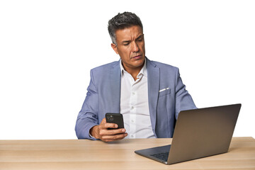 Serious middle-aged Hispanic man on phone while working on laptop