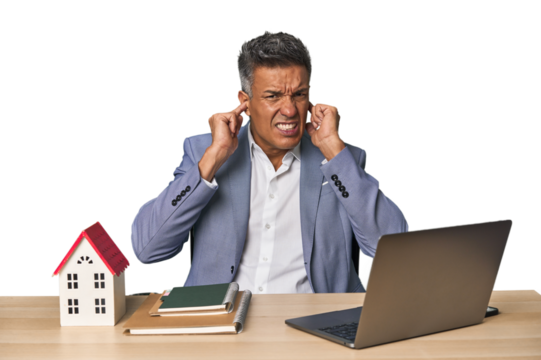 Middle-aged Hispanic businessman with real estate theme covering ears with hands.