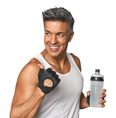 Gym-ready Hispanic man with water bottle points with thumb finger away, laughing and carefree.