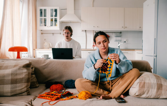 Young woman knitting crocheting with colored yarn granny square while watching online tutorial on phone. And young man working on laptop from homeWoman watching needlework lessons, home hobbies