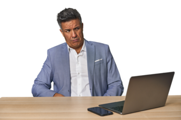 Elegant businessman at desk with laptop confused, feels doubtful and unsure.