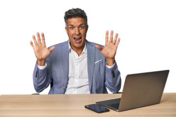 Elegant businessman at desk with laptop receiving a pleasant surprise, excited and raising hands.