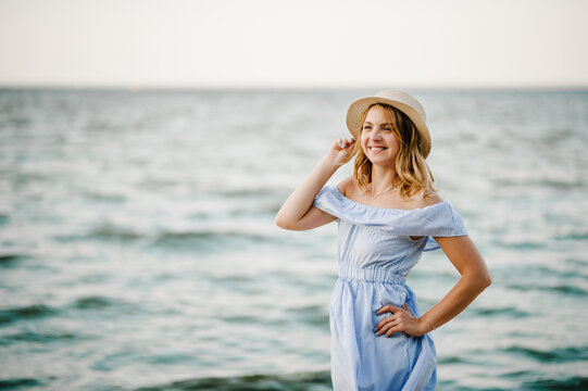 Young attractive smiling blond woman in straw hat joyfully walks near sea. Happy beautiful girl standing on beach ocean and enjoying sunny summer day on vacation. Portrait of stylish female closeup.