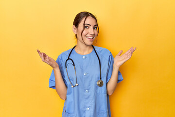 Young nurse in a yellow studio receiving a pleasant surprise, excited and raising hands.