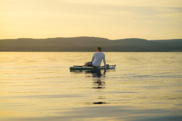 A man sits with his back facing on a paddleboard, relaxing and enjoying the sunset and vacation.