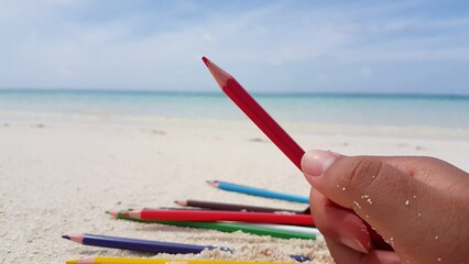 Shallow focus of a hand holding a red pencil with blur background of a sandy beach and blue water