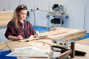 Woman furniture maker. Girl carpenter measures size of board. Furniture shop employee with tape measure. Carpenter near construction drawings. Woman furnisher at work. Carpenter at table with boards - obrazy, fototapety, plakaty