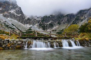 Photo of the rocky peaks of the High Tatras above the cascade flowing from the glacial lake under the pedestrian bridge during a cloudy day - obrazy, fototapety, plakaty