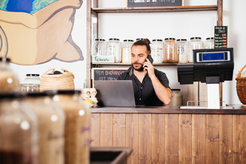 Storekeeper Managing Business Affairs on Laptop While Conversing on Phone at Grocery Counter