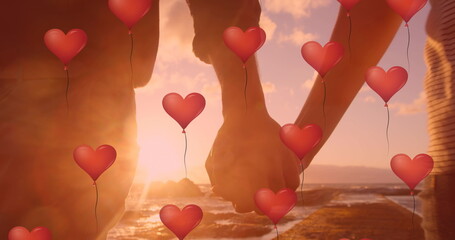 Image of red heart balloons floating over mid section of couple in love holding hands by seaside