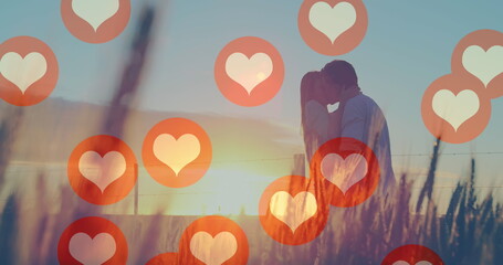 Image of red heart icons floating over mid section of couple in love kissing at sunset