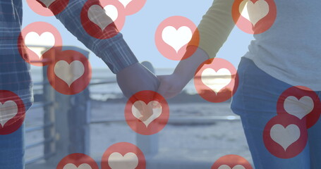 Image of red heart icons floating over mid section of couple in love holding hands by seaside - obrazy, fototapety, plakaty