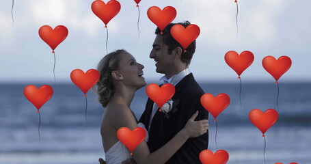 Fototapeta premium Image of multiple red heart balloons floating over newly married couple embracing on beach