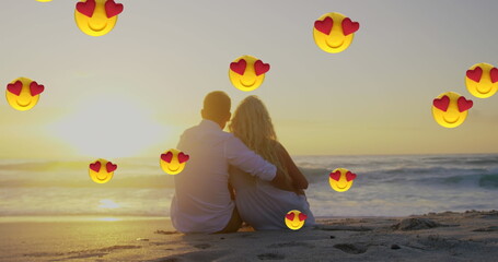 Image of multiple heart eyes face emoji floating against couple in love embracing sitting on beach